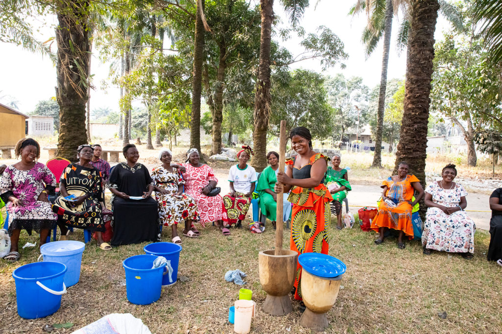Une femme africaine tenant un bâton entrain de moudre. Autour d'elles, des femmes africaines et la regardent