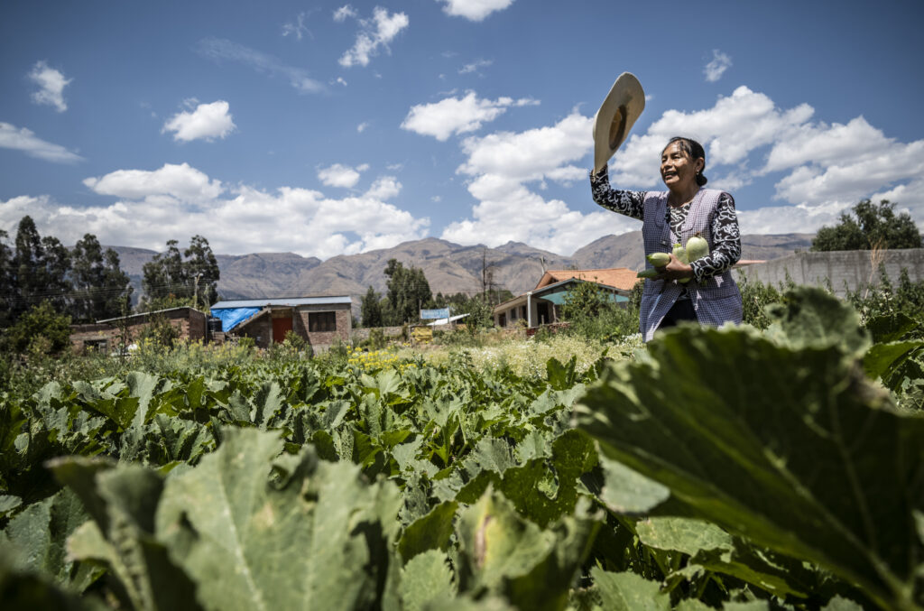 Paysanne bolivienne dans son champ qui tient des courgettes dans ses bras et salue avec son chapeau de l'autre main